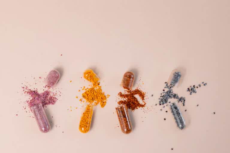 Pill-shaped capsules with colored powder spills: pink, orange, brown, and blue scattered on white background.