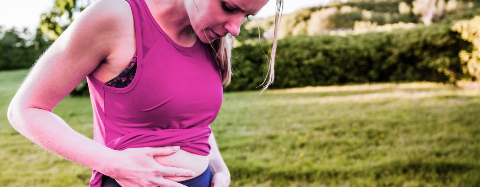 Woman in purple tank top clutching her abdomen in outdoor park, looking pained while standing on grass.