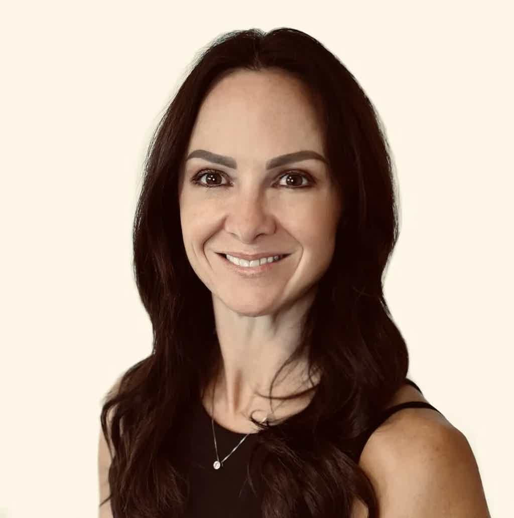 Smiling woman with long dark hair wearing a black top and a pendant necklace, neutral background.