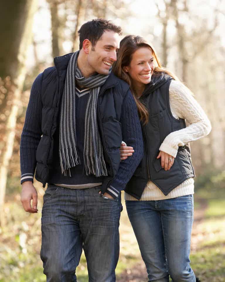 A smiling couple walks arm-in-arm through a sunlit forest, wearing jackets and scarves.