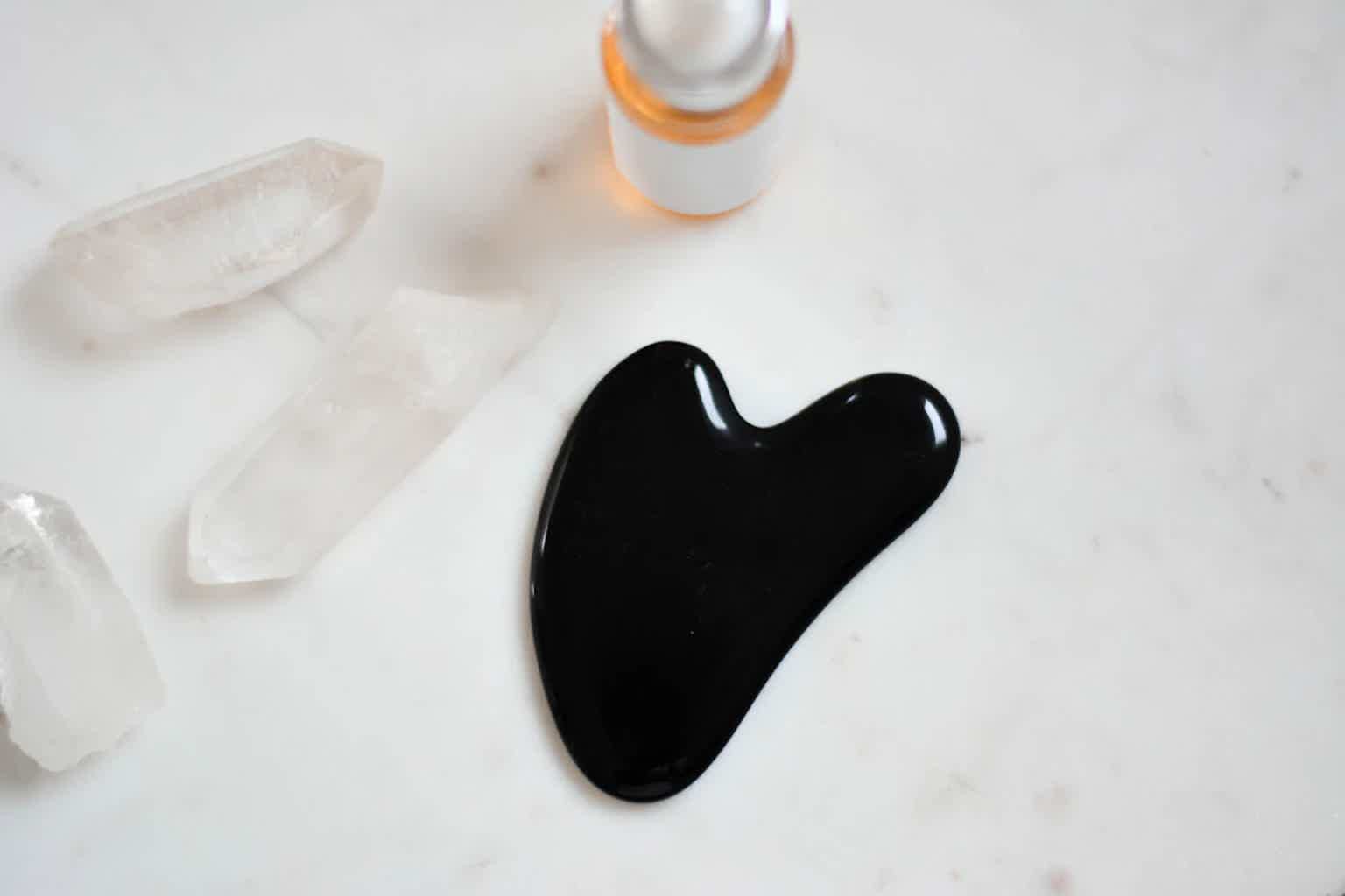 Black gua sha stone with curved edge on white marble, with quartz chunks and small skincare bottle above.