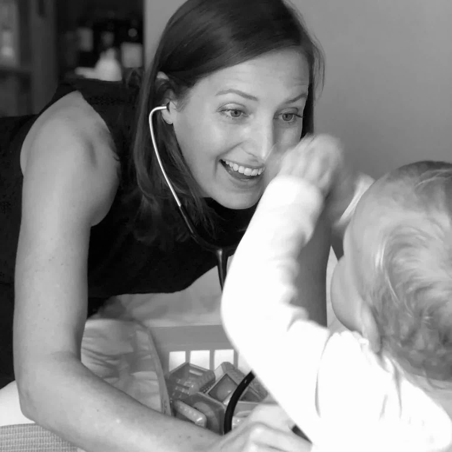 A smiling woman in a black top uses a stethoscope while playfully interacting with a baby in a cozy setting.