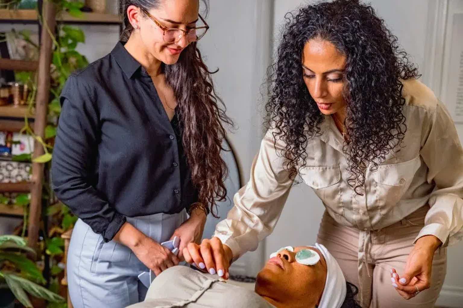 Two women in work uniforms gently treating a client with a facial mask, seated in a spa treatment room.