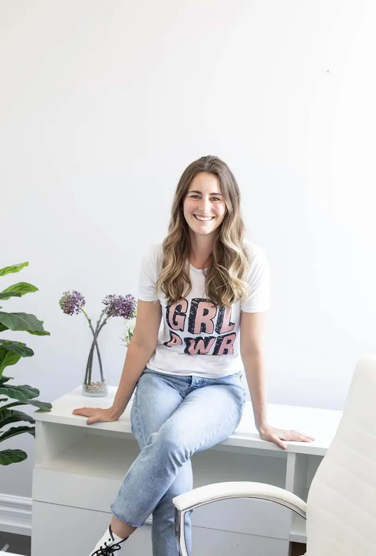 Smiling woman seated on a white office chair, wearing a “GRL PWR” T-shirt and light jeans, with plants nearby.