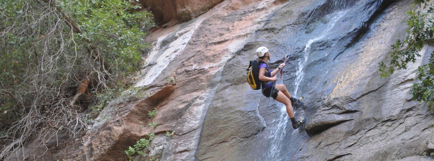 Hiker with backpack climbs a steep rock face beside a small waterfall in a green canyon.