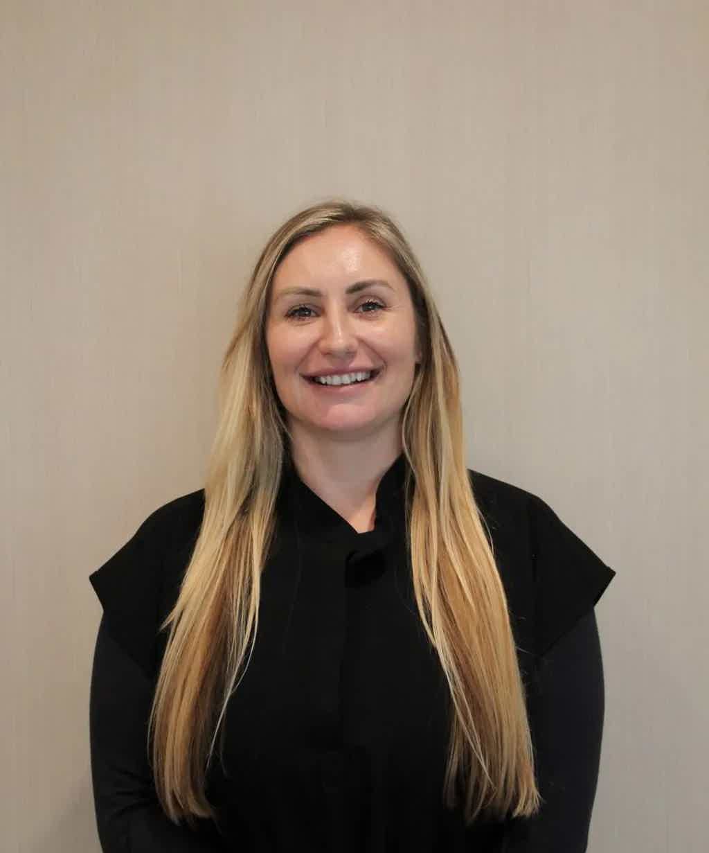 Smiling woman with long blonde hair wearing a black top stands against a light beige wall.