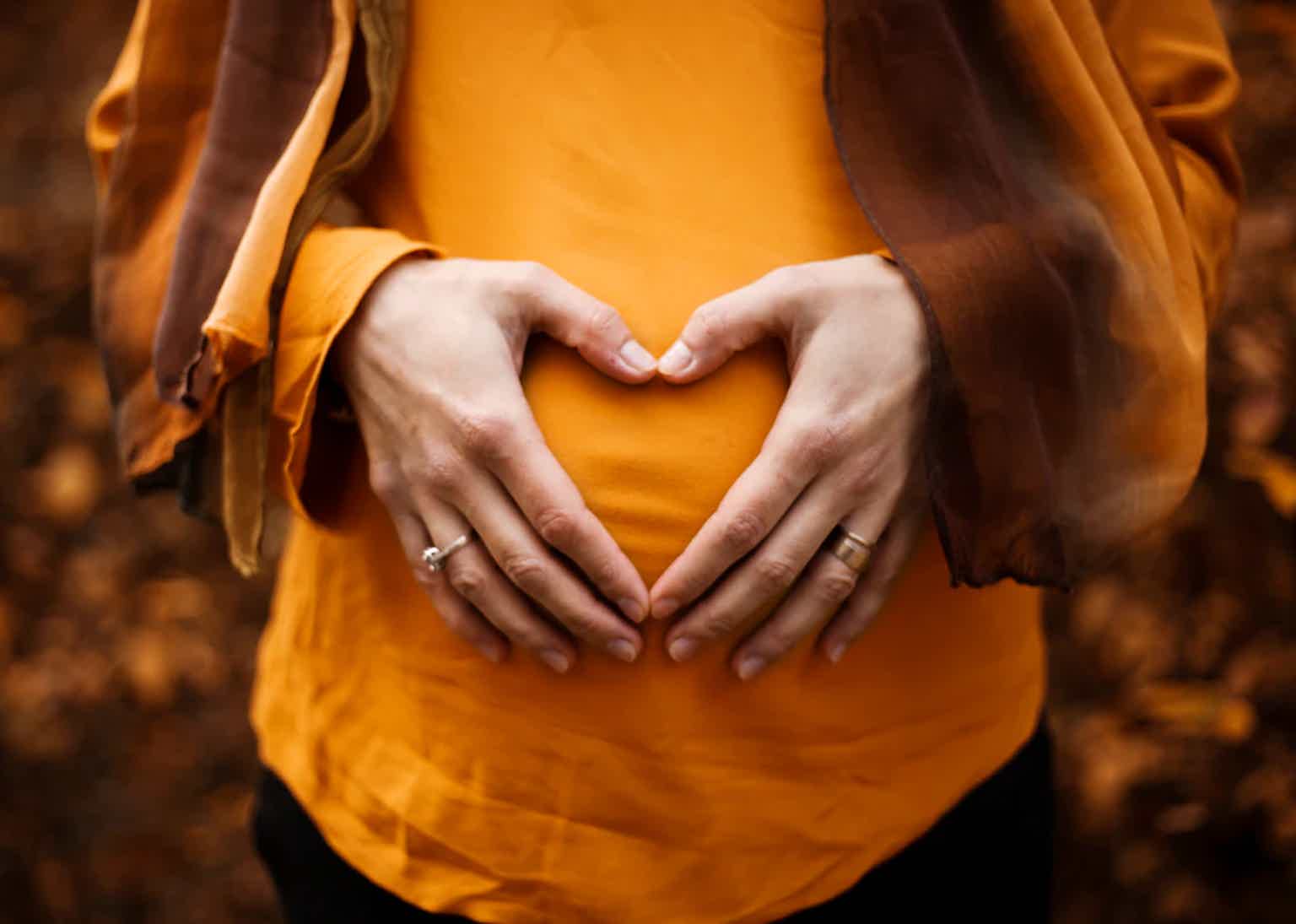 Pregnant belly framed by hands forming a heart shape, warm orange tones.