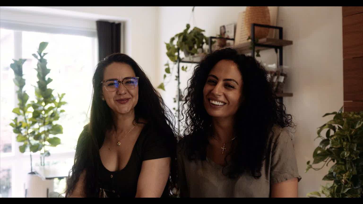 Two women with curly and straight dark hair smile at the camera in a bright indoor room with plants.