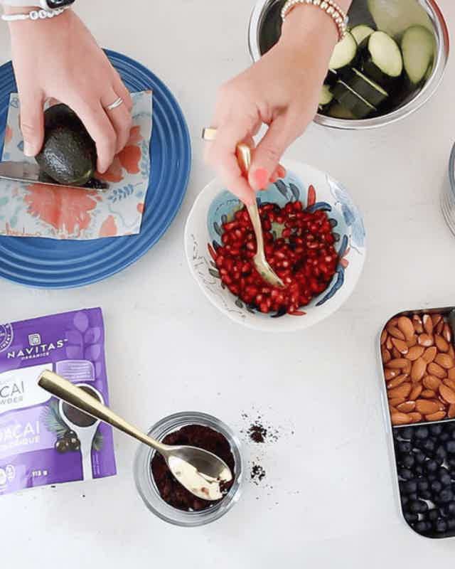 Hands prep ingredients: slicing avocado, pomegranate seeds in bowl, almonds, blueberries, cucumber slices in bowl, purple pac