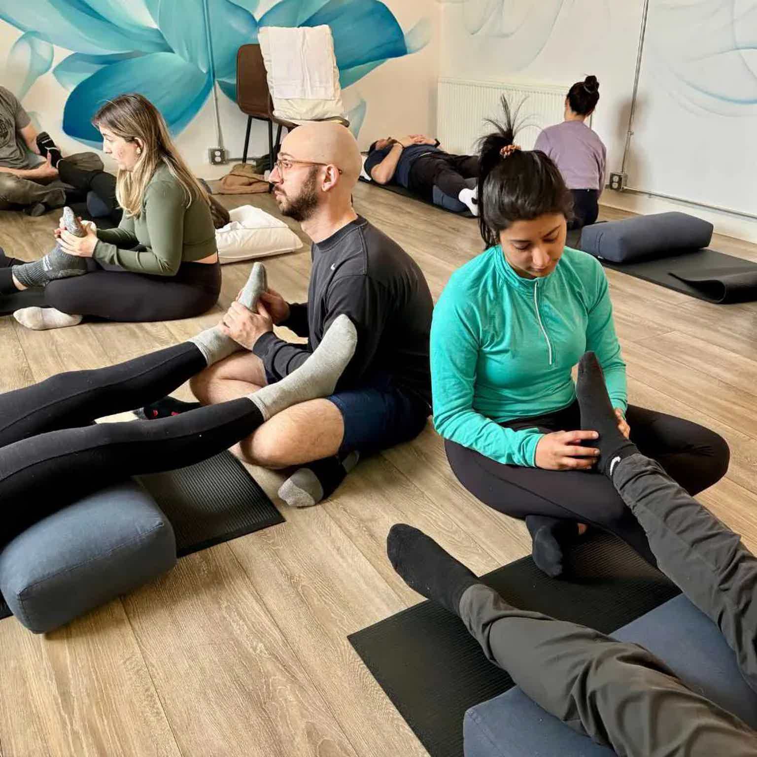 People in a stretching/yoga class seated and lying on mats in a studio with foam rollers and towels.