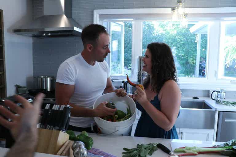 Two people cooking together in a bright kitchen, chopping and tossing veggies at the counter while smiling.