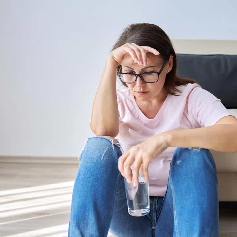 Woman sitting on floor with head in hand, holding a glass of water, looks distressed.