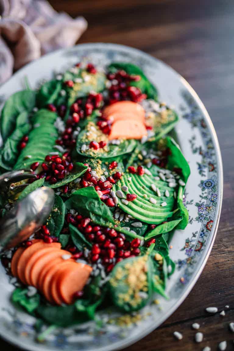 Fresh salad with avocado slices, spinach, cucumber, pomegranate seeds, and sesame on a floral plate.
