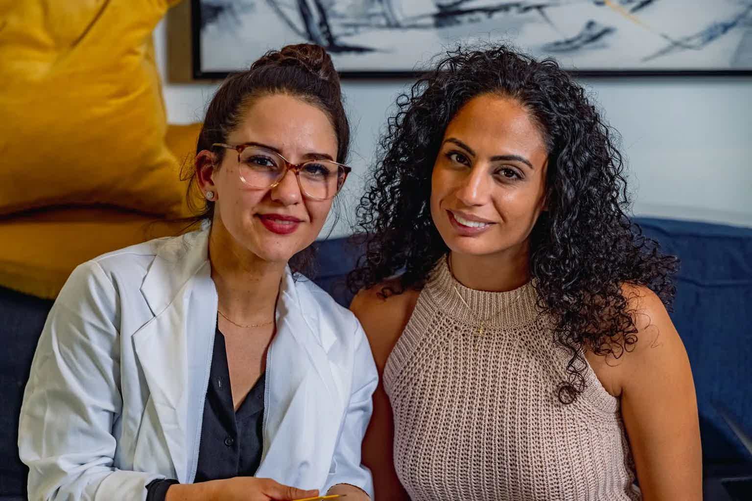 Two women seated side by side, one in a white lab coat with glasses, the other with curly hair and a sleeveless sweater.