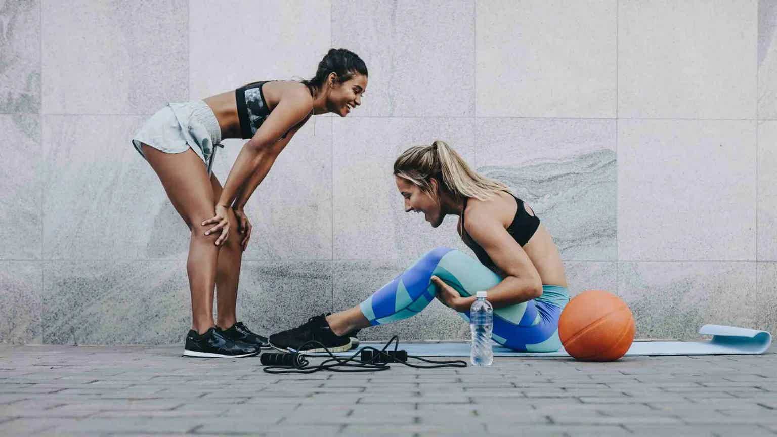 Two women stretching outdoors on a brick patio; one kneels while holding a leg, yoga mat and ball nearby.