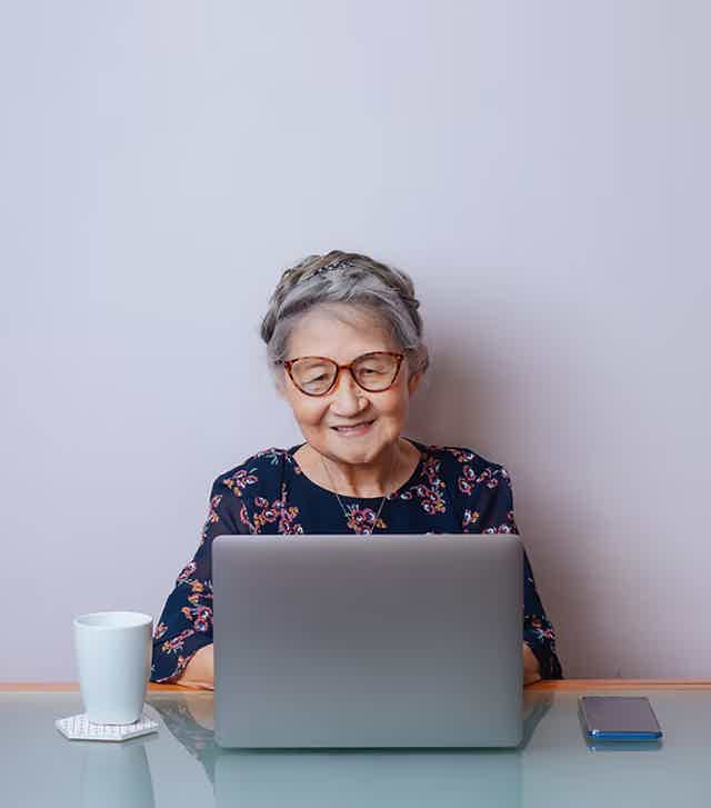 Senior woman with gray hair and glasses sits at a desk using a laptop, with a mug and phone nearby.