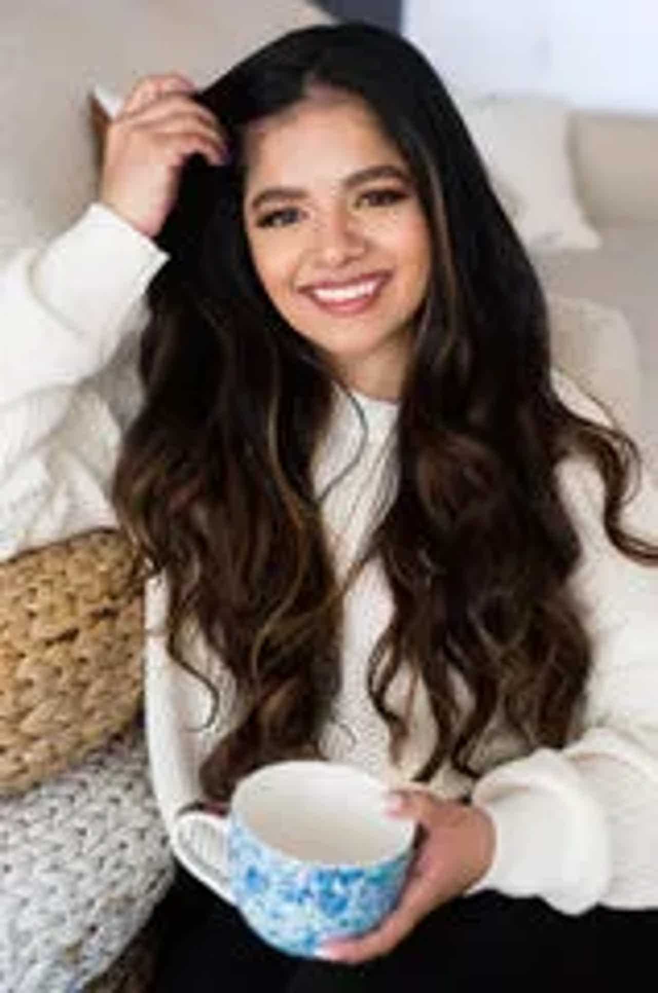 Smiling woman with long dark hair, white sweater, seated on pink chair, plant beside her in a bright room.