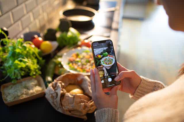 Person holding smartphone capturing a colorful market food display with herbs, eggs, and mushrooms in a shop aisle.