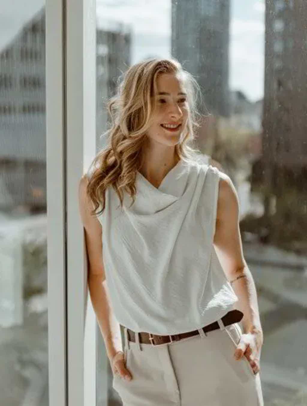 Woman in white sleeveless top and light pants with belt, standing by window, smiling in urban setting.