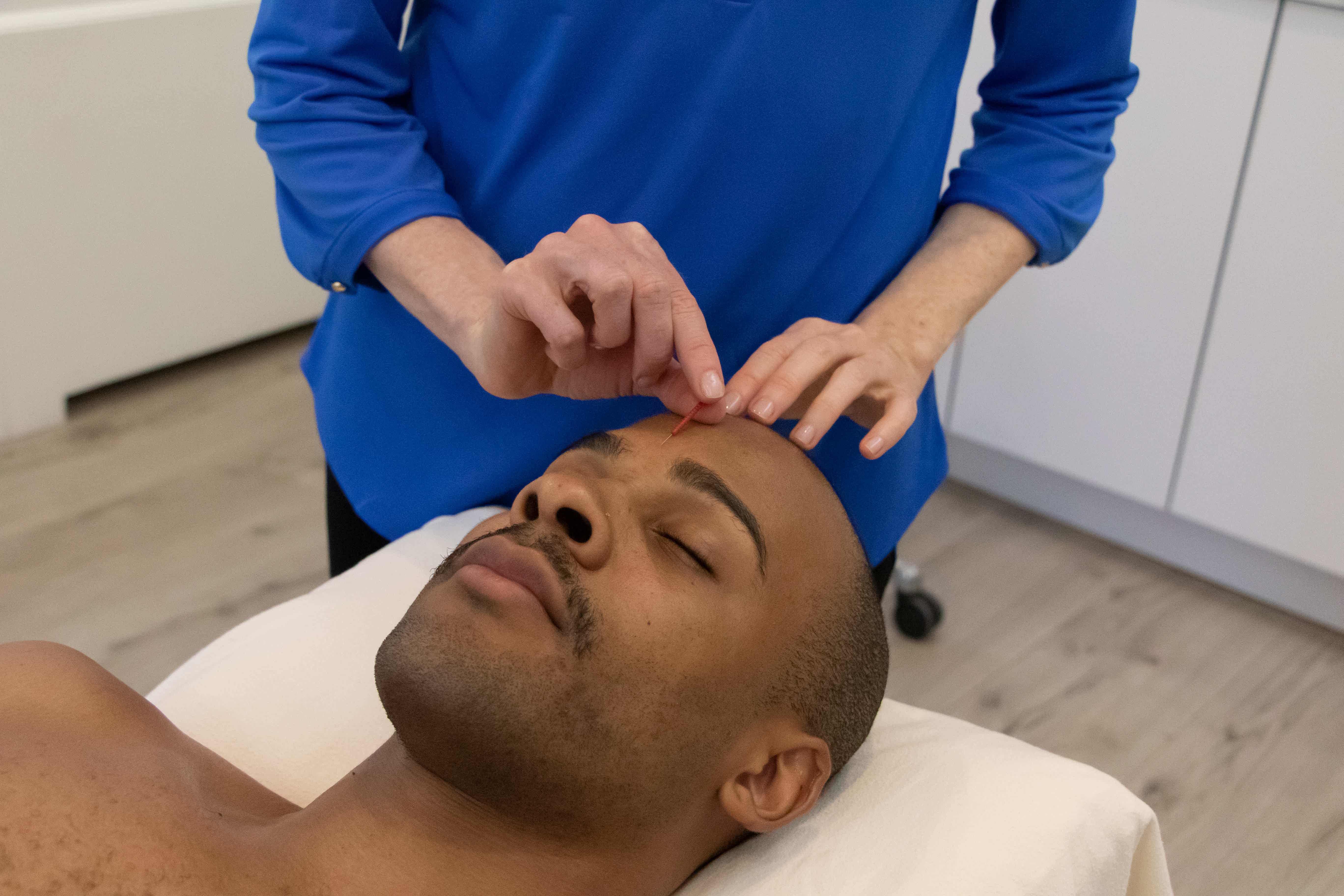 Practitioner in blue places an acupuncture needle as the patient relaxes in a clinic room