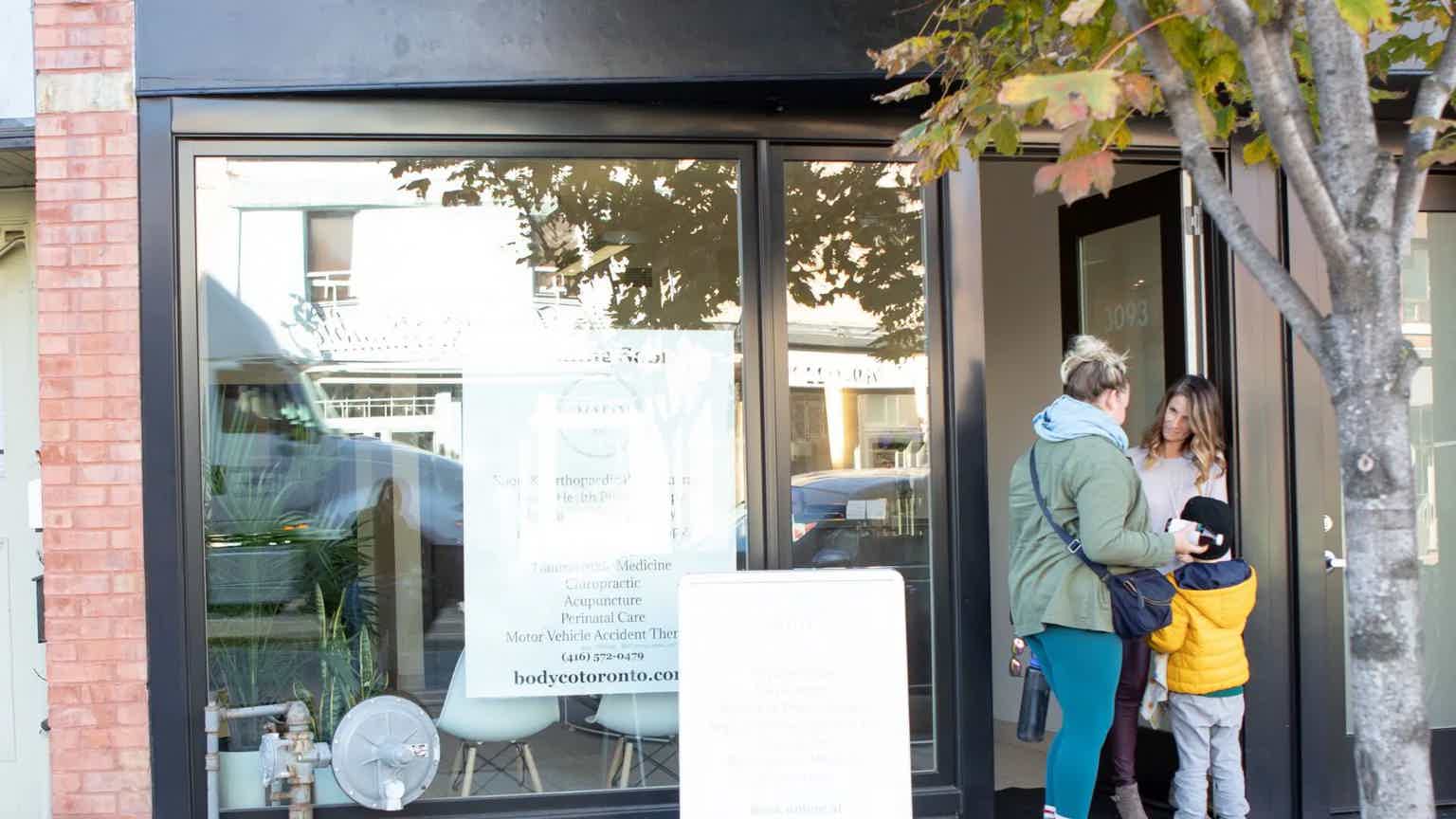 People walking past building entrance with glass door and storefront sign; tree branches and brick columns nearby.