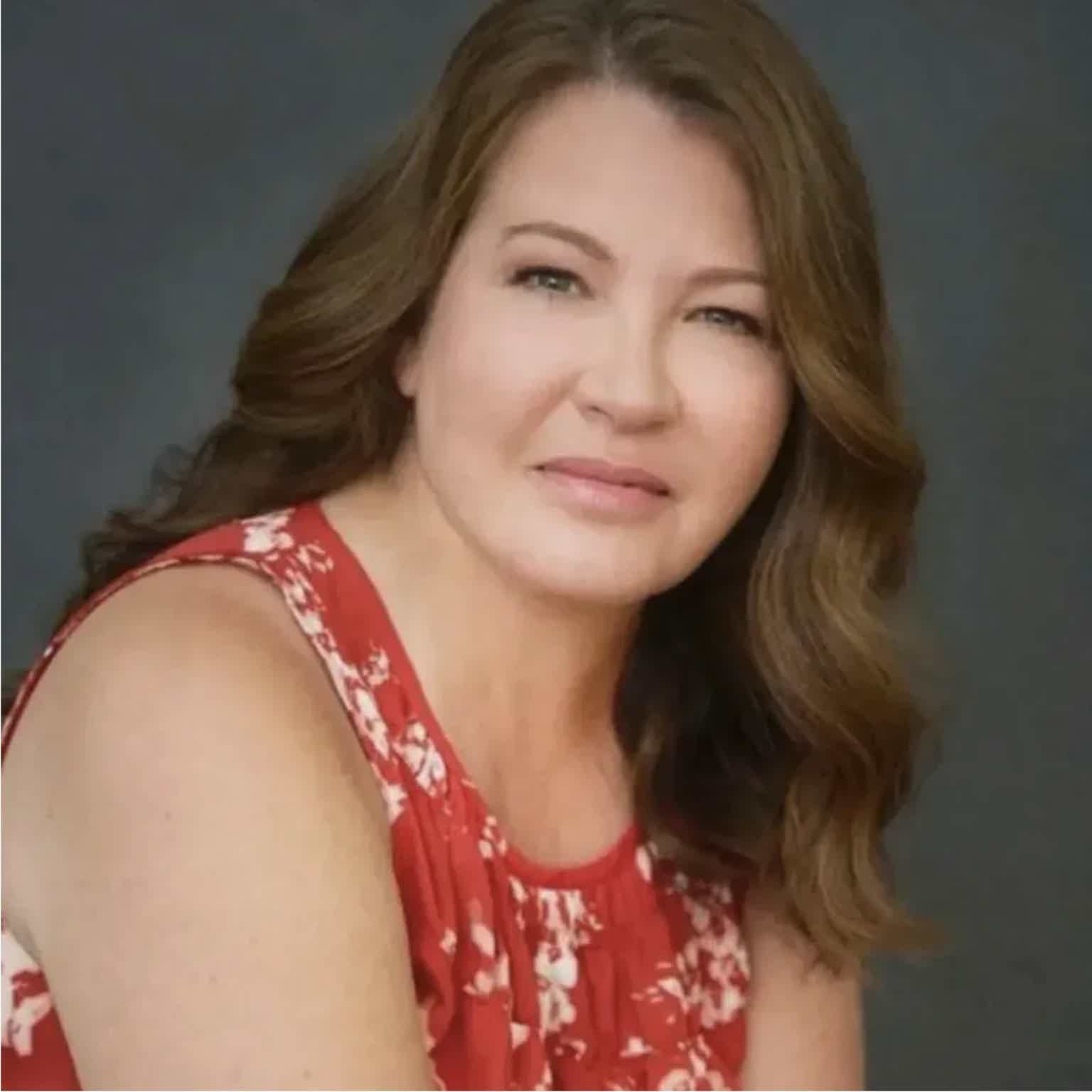 Smiling woman with shoulder-length auburn hair wearing a red sleeveless floral dress against a neutral gray background