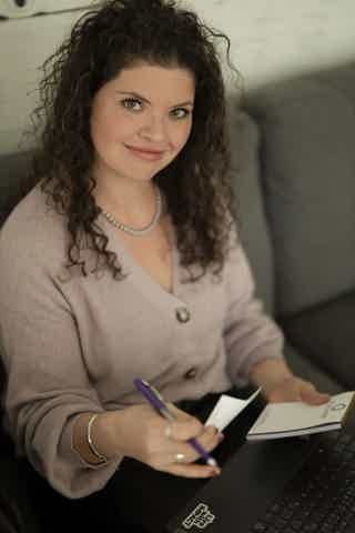 Woman with curly hair sits on a couch, writes on notepad and uses a laptop, wearing a pink cardigan and pearl necklace.