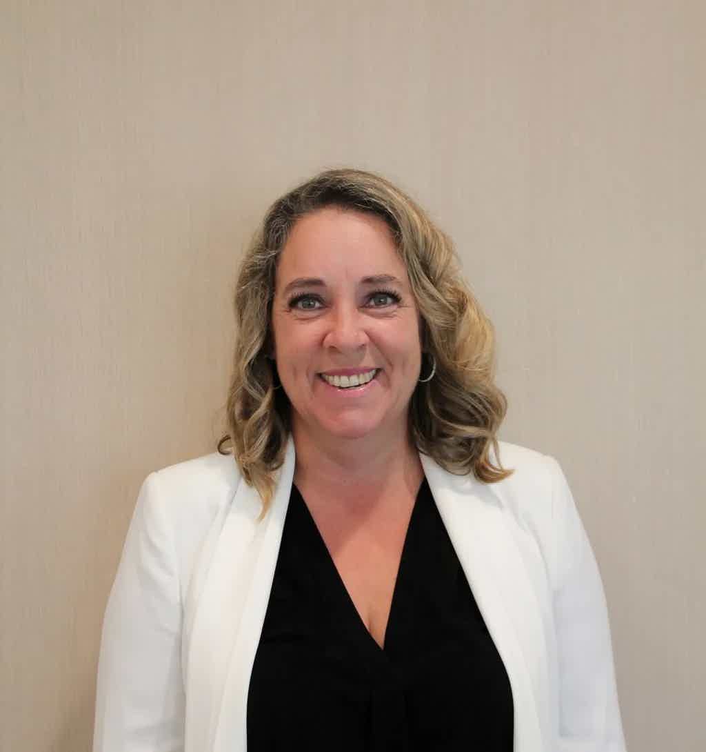 Smiling woman with shoulder-length wavy hair in a white blazer over a black top, neutral background.