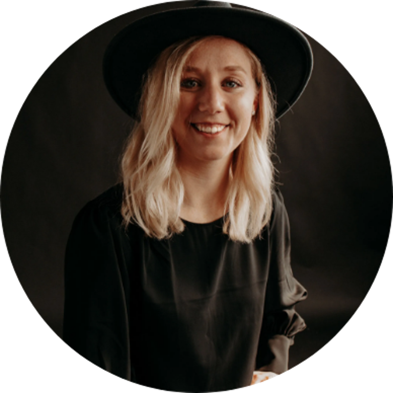 Smiling woman with blonde hair wearing a black wide-brim hat and black top, sitting against a dark backdrop.