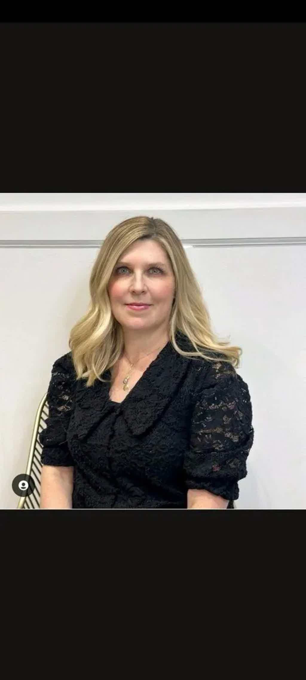 Blonde woman in black lace top sits against white wall, slight smile, necklace visible.