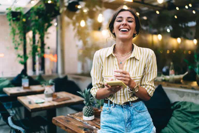 Young woman laughing while holding a phone at an outdoor cafe with string lights and plants.