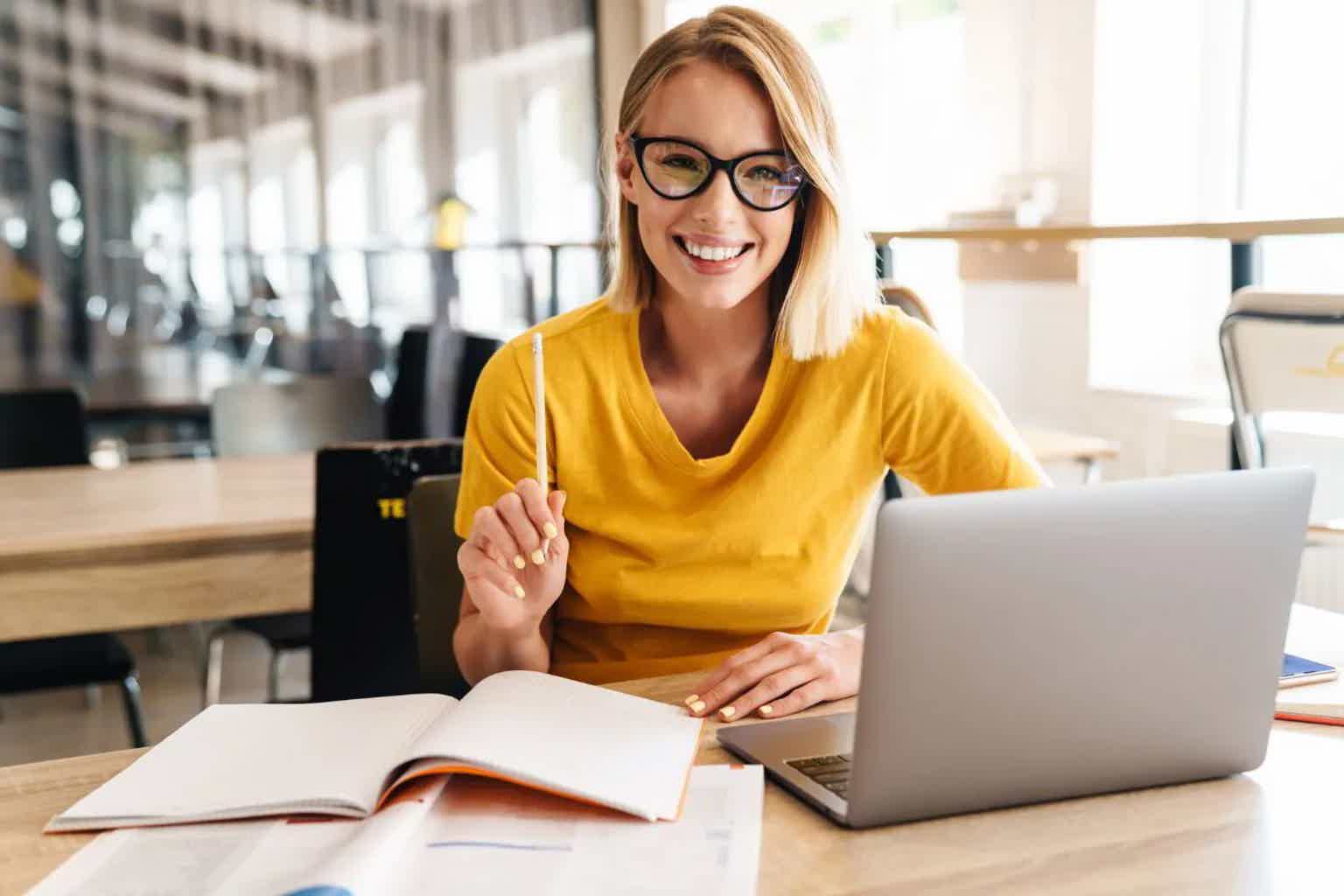 Smiling woman with glasses in yellow top working at a café table with open laptop and an open book.