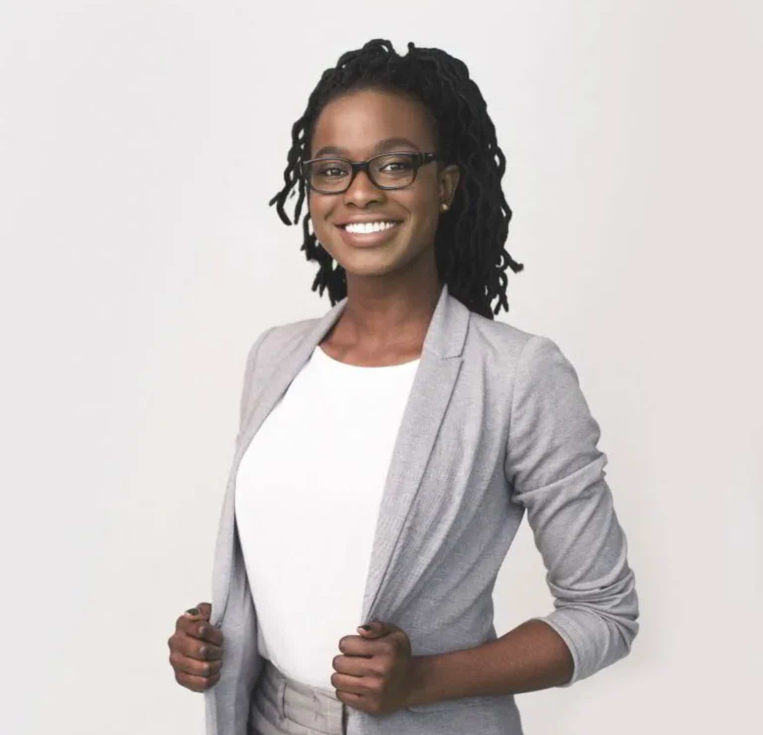 Smiling woman with glasses and dreadlocks wearing a white top and light gray cardigan, standing against a plain background.