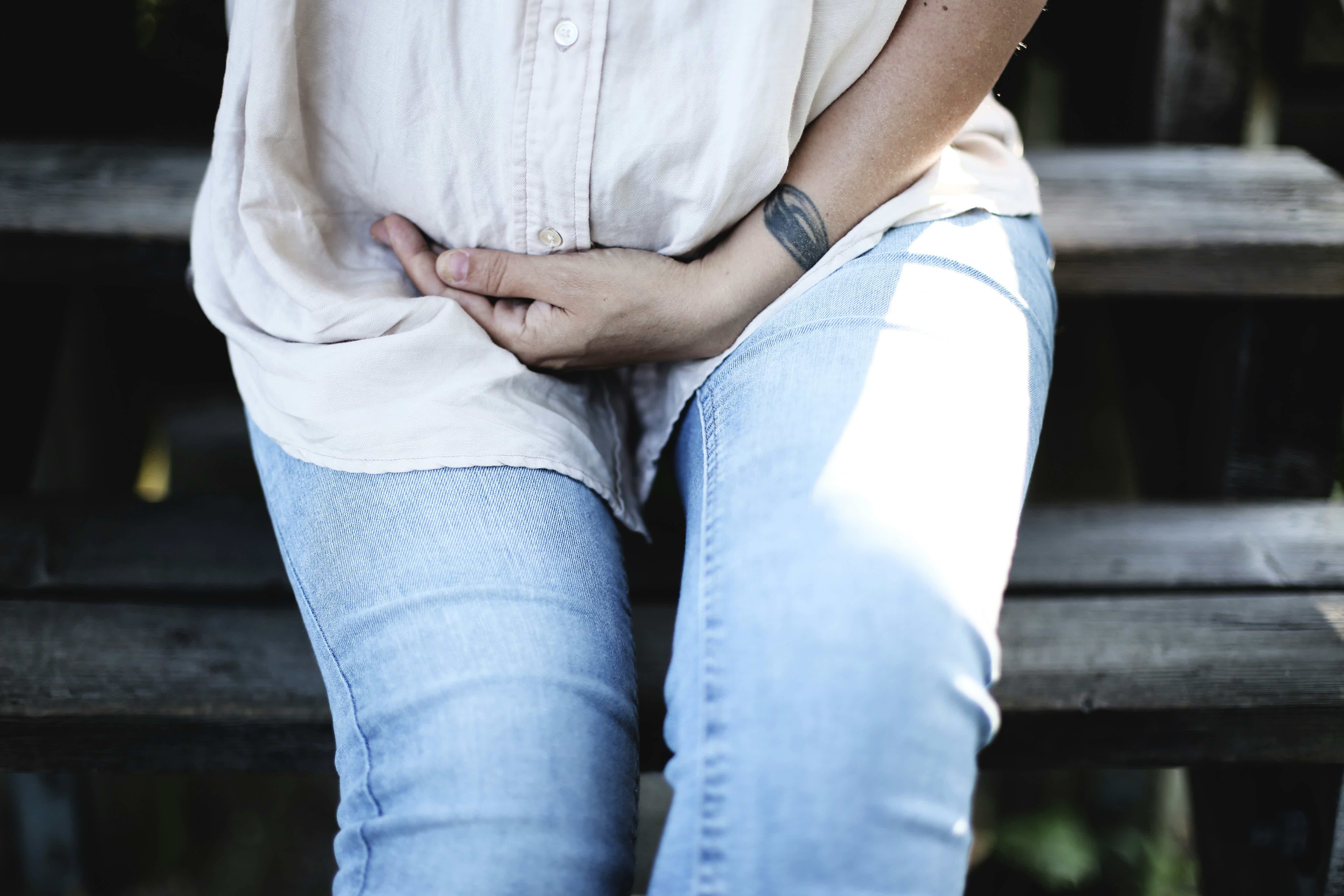 Person seated on steps hugging knees, wearing a white shirt and light blue jeans; arm tattoo visible.