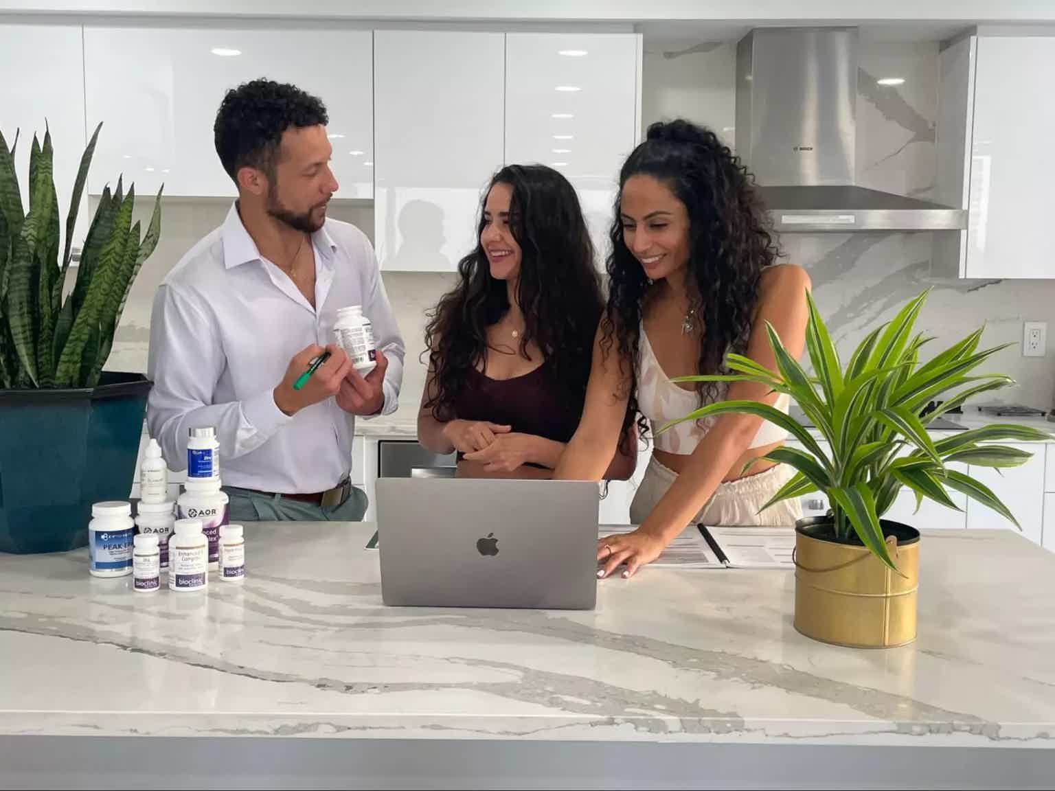 Three people smiling and discussing at a kitchen counter with an open laptop, plant in foreground, and skincare bottles.