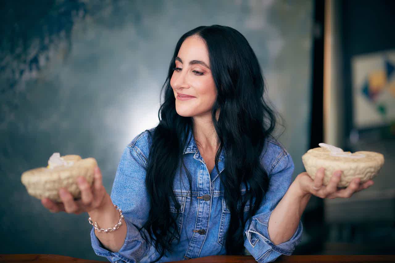Woman with long dark hair in denim shirt holding two bowls of dessert, looking thoughtfully to the side.