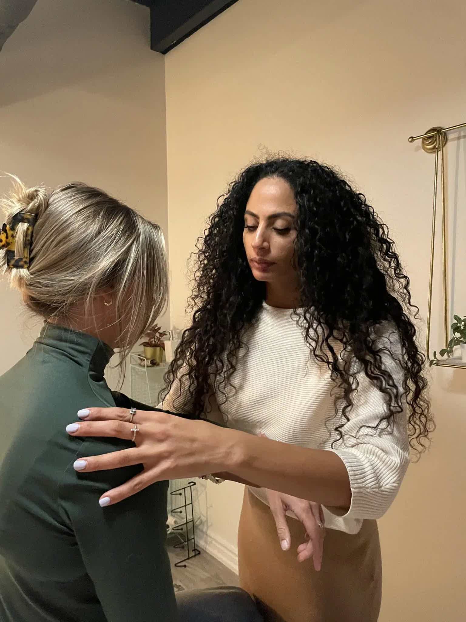 Woman with long curly dark hair standing while another person styles her hair in a salon, indoor mirror backdrop.