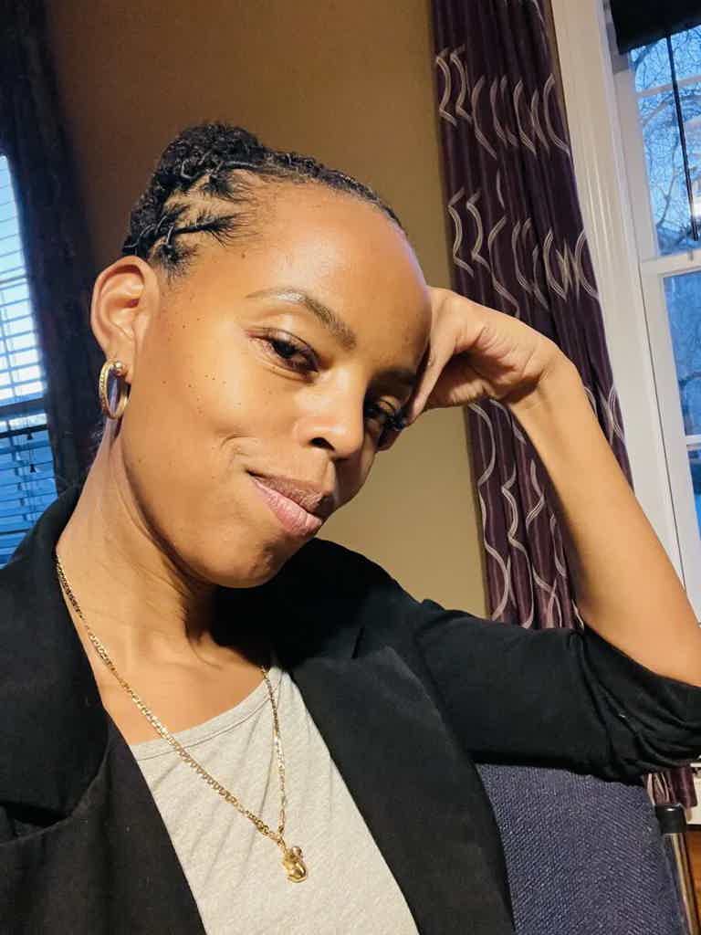 Woman with braided hair, smiling slightly, resting cheek on hand, wearing black blazer and gold jewelry.