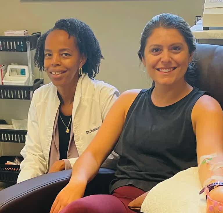 Two smiling women in a clinic: one in white coat, the other in dark tank top, arm IV line, medical office setting.