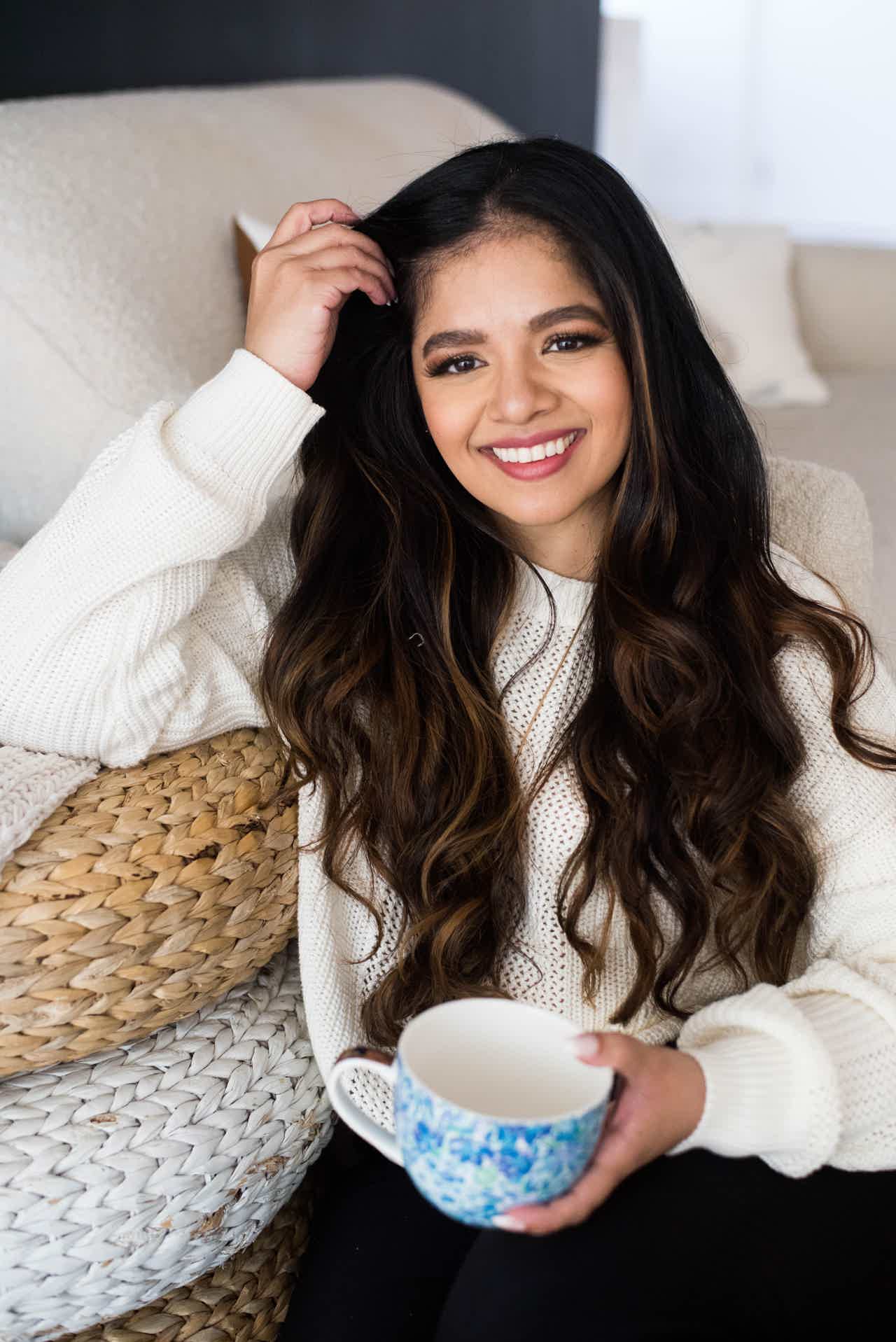 Young woman with long dark hair smiling, holding a blue mug, sitting on a couch.