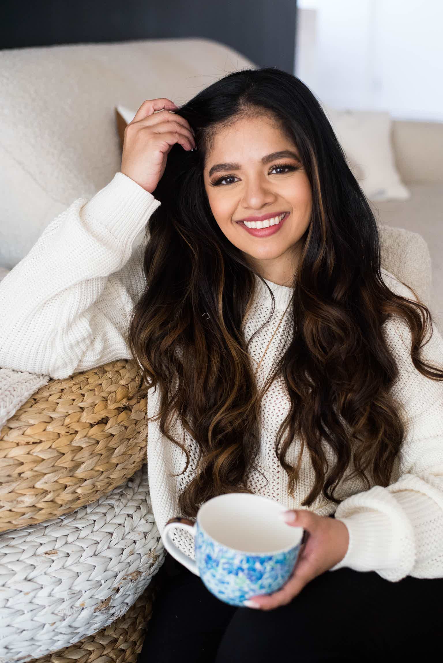 Young woman with long dark hair smiling, holding a blue mug, sitting on a couch.