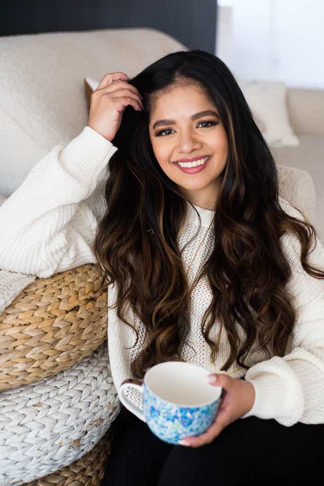 Young woman with long dark hair smiling, holding a blue mug, sitting on a couch.