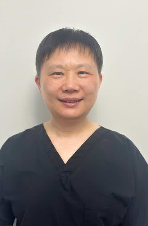 Smiling woman in light sweater and black leggings seated barefoot on a wooden chair, posing against a white background.