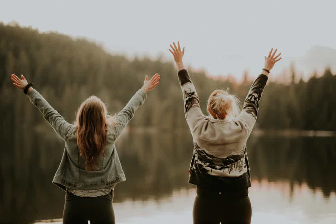 Two people stand by a lake with arms raised, facing the water and forest beyond.