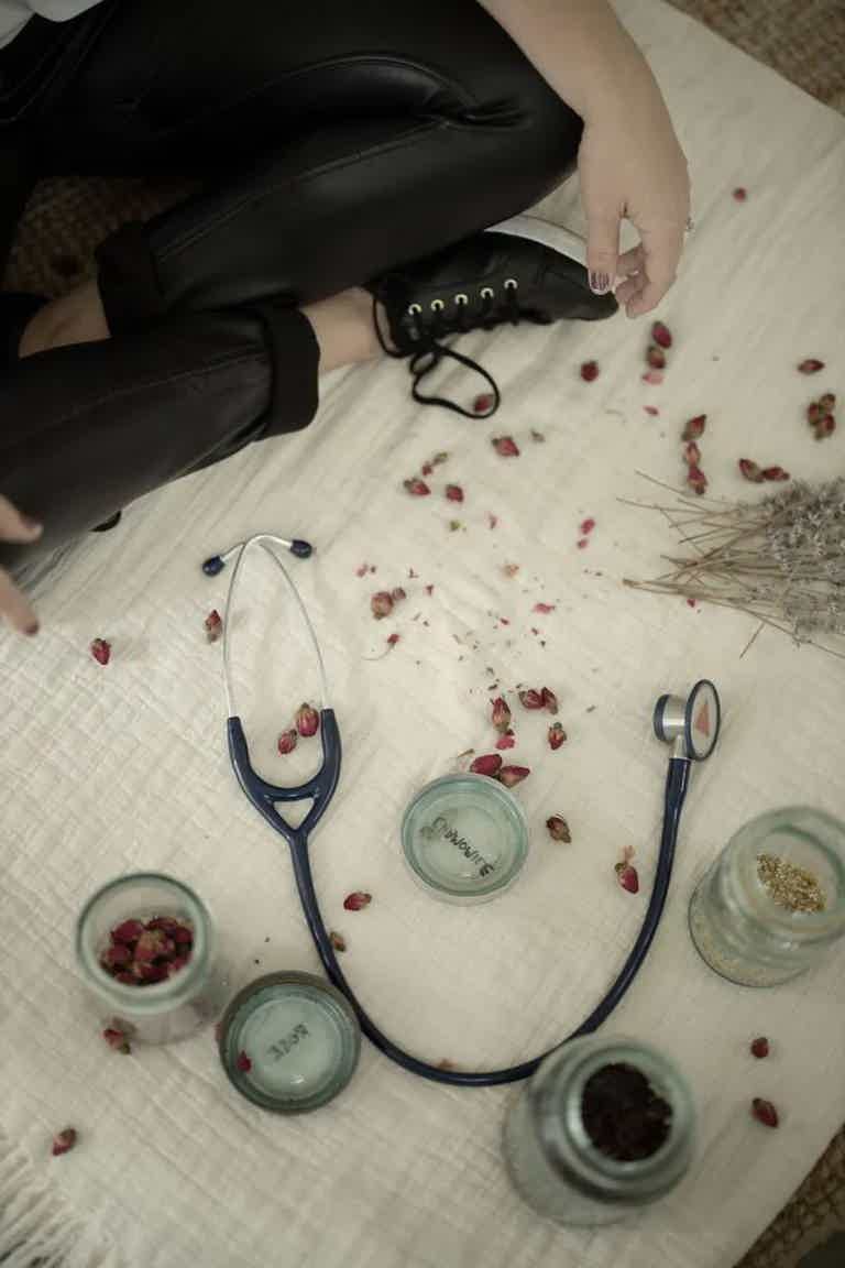 Person seated on floor with medical tools: a stethoscope, glass jars, dried rose petals, and a hand near a boot.