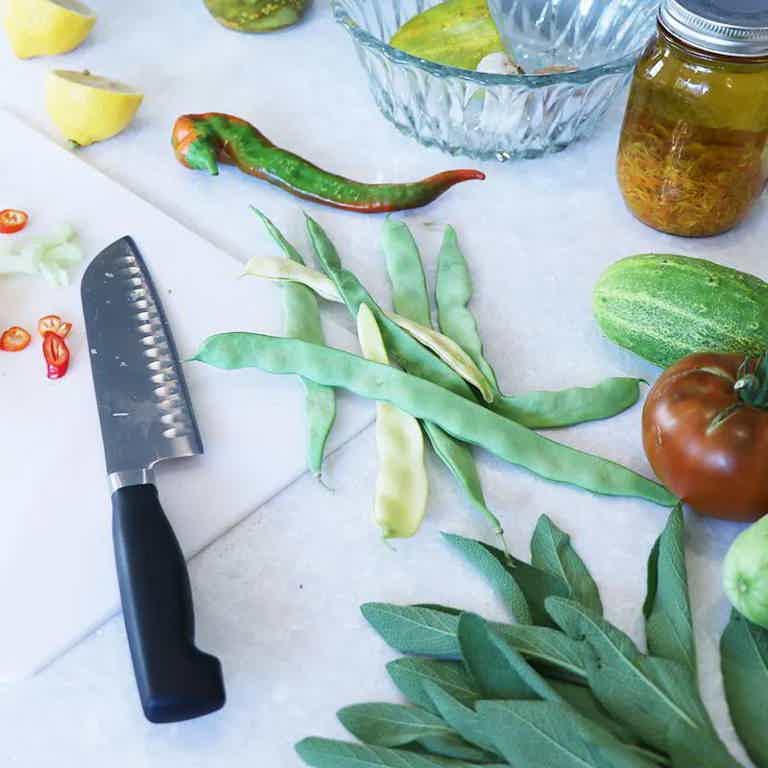 Assorted fresh herbs, beans, peppers, and vegetables with a knife on a cutting board and jar in the background.