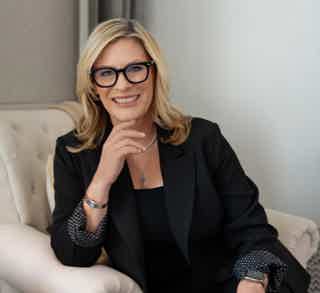 Blonde woman in black blazer and glasses sits on beige chair, smiling with hand on chin.