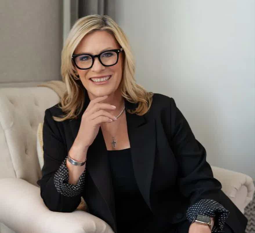Blonde woman in black blazer and glasses sits on beige chair, smiling with hand on chin.
