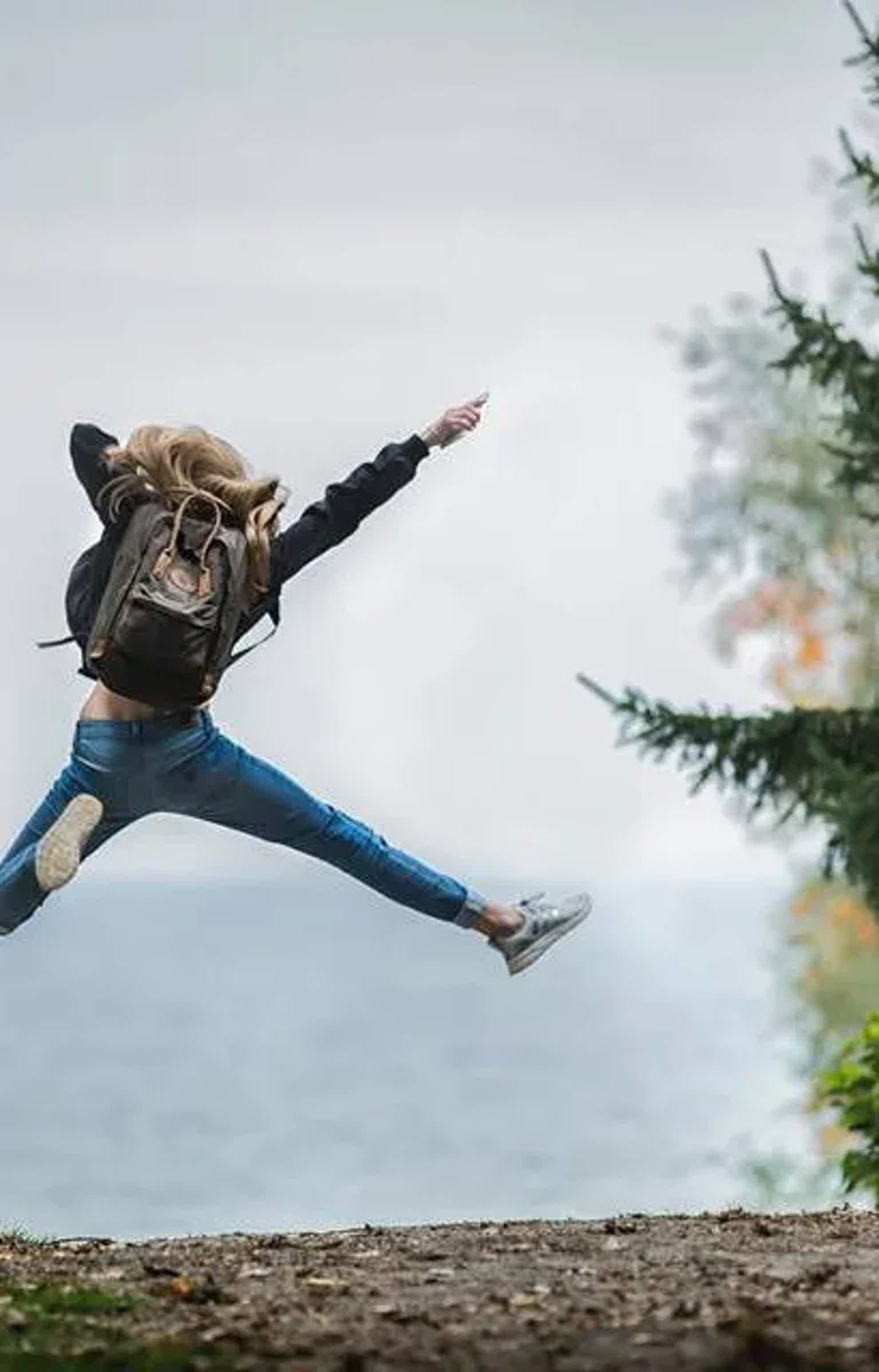 Person mid-jump on a gravel path, backpack on back, arms outstretched, with trees blurred in background.