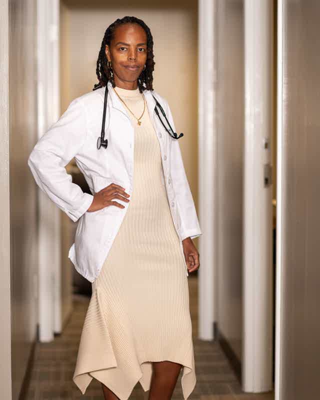 Female doctor in beige dress and white coat with stethoscope in hallway, hands on hips, confident pose.